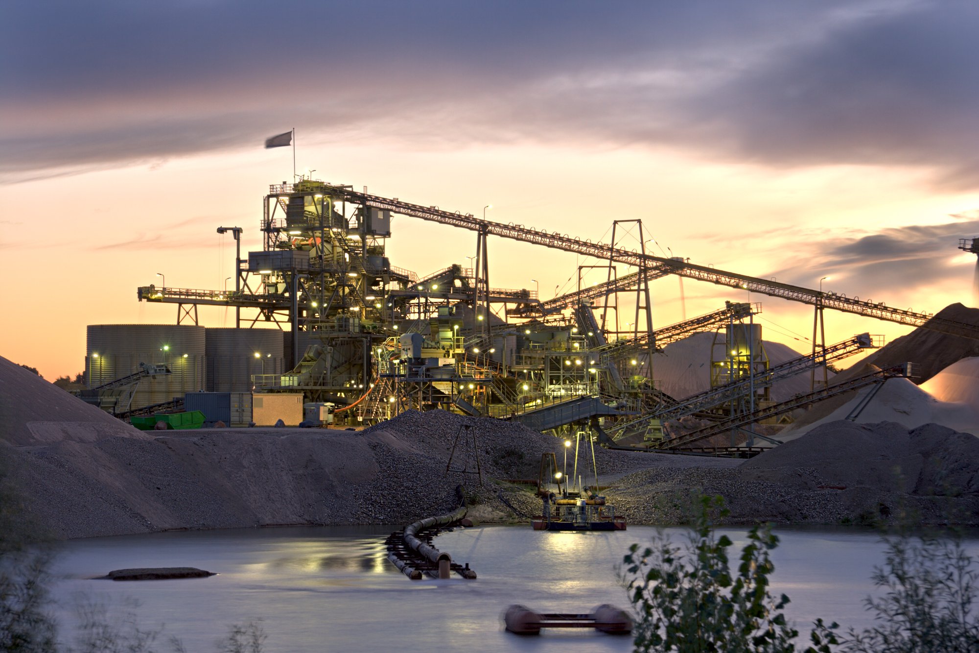 Aggregate processing plant lit at dusk beside a water pond